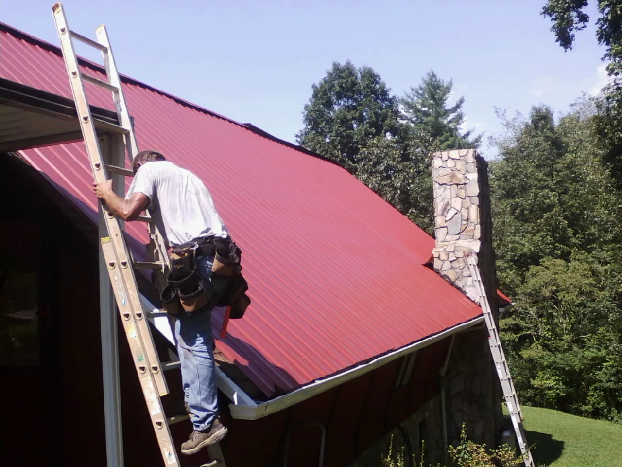 Roofer on a ladder beside a red metal roof and stone chimney — Soffit & Fascia Repair by Roofing Brothers in Belfast